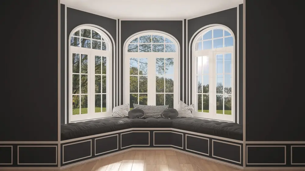 A cozy bay window nook with black and white cushioned seating and striped pillows, surrounded by large arched bay windows letting in natural light and offering views of the lush home exterior and green trees outside.
