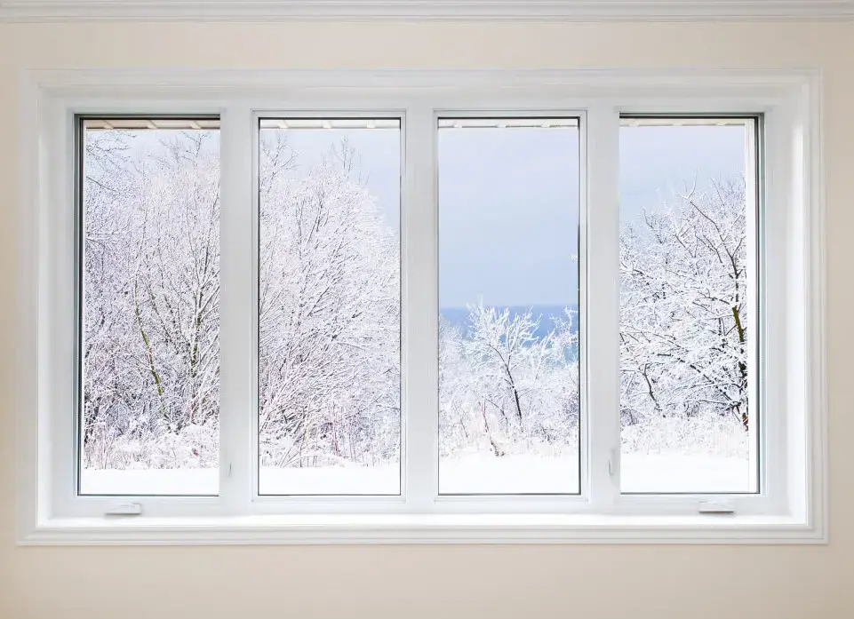 Four-panel energy efficient windows in MA frame a snowy landscape with bare, snow-covered trees. Set against a pale beige wall, the view features a light blue, partly cloudy sky.