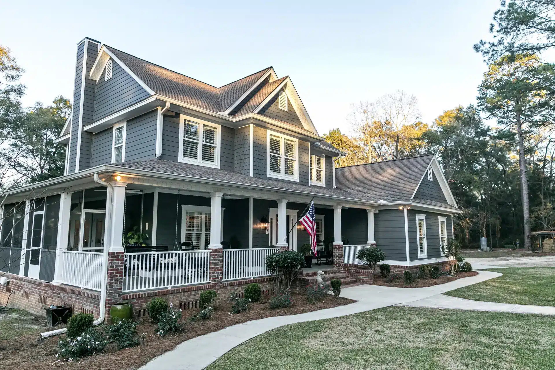 A two-story gray house with white trim and a wraparound porch, displaying an American flag, evokes the charm of a Farmers Almanac New England homestead, nestled among trees and landscaped yard with a curved sidewalk.