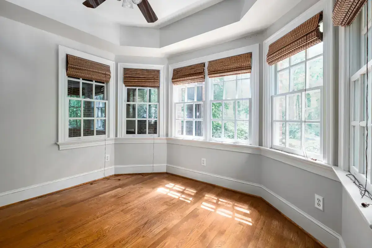 A bright, empty room with hardwood floors, light gray walls, and multiple large windows with woven shades—the best windows for natural light. Sunlight streams in, casting patterns on the floor. A ceiling fan is visible above.