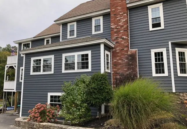 Two-story house with dark gray siding, white trim, and a red brick chimney. Several windows—recently upgraded with affordable window installation—and shrubs are visible in the landscaped yard. A stone foundation supports the house.