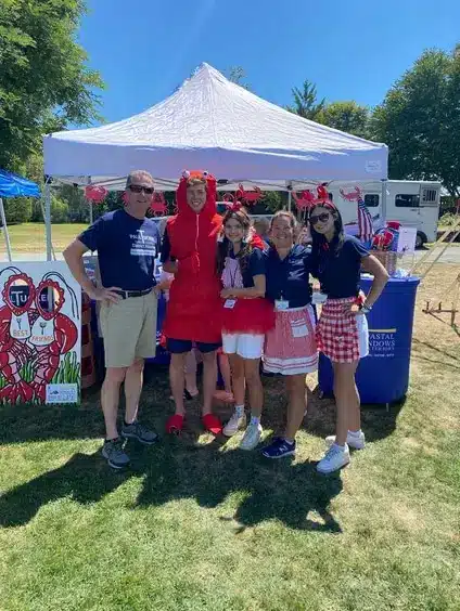 Five people pose in front of a white tent at an outdoor event, one dressed as a red lobster. The others wear matching navy shirts and summer attire, their smiles bright as they discuss the Window Replacement Checklist under a sunny sky.