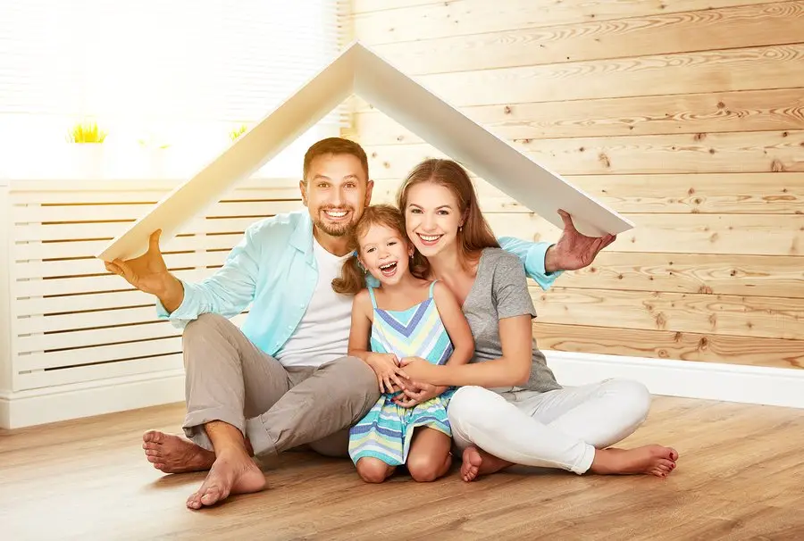 A smiling family of three sits on a wooden floor, holding a large white board over their heads like a roof, symbolizing protection, a happy home, and the security that comes with knowing your roof replacement FAQ.