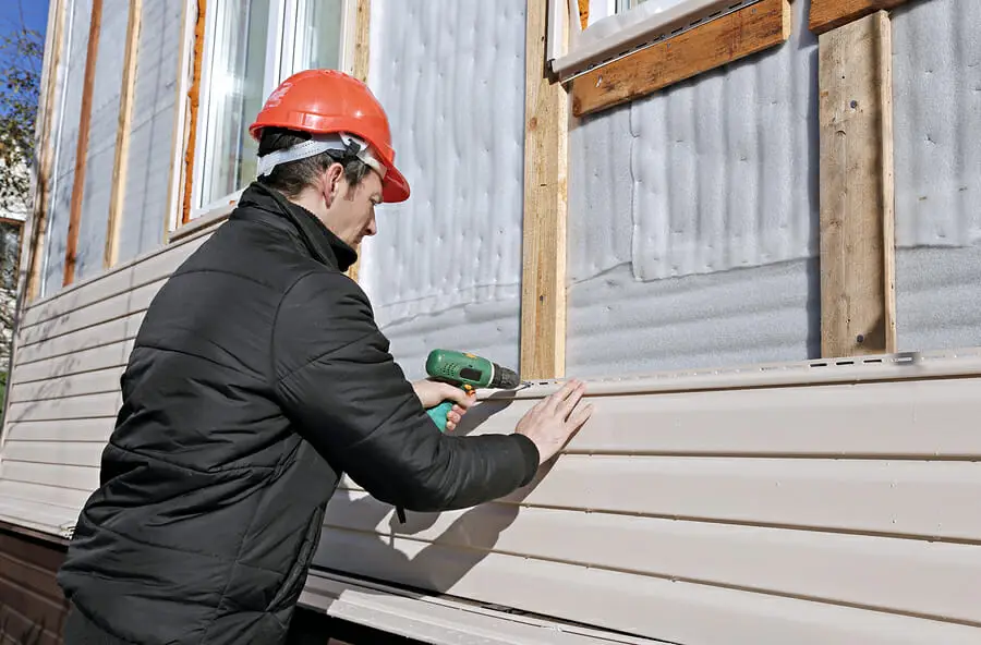 A person wearing a red hard hat and black jacket uses a power drill to install beige vinyl siding in MA on the exterior of a house under construction.