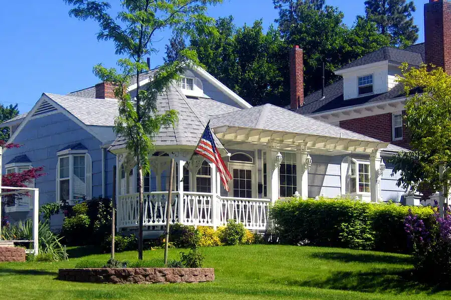 A light blue house with white porch and railing exudes Americana curb appeal, featuring an American flag out front, lush green grass, bushes, trees, and landscaped flower beds beneath a clear blue sky.