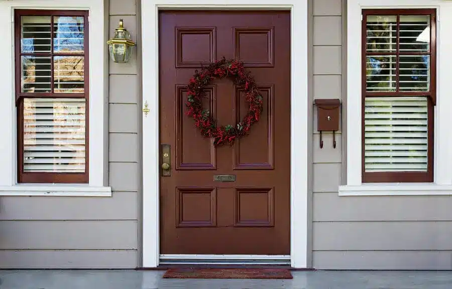 A brown entry door with a berry wreath is centered between two windows with white shutters. A brass light fixture sits on the left, and a brown mailbox is mounted on the right. The light gray exterior wall completes the charming look.