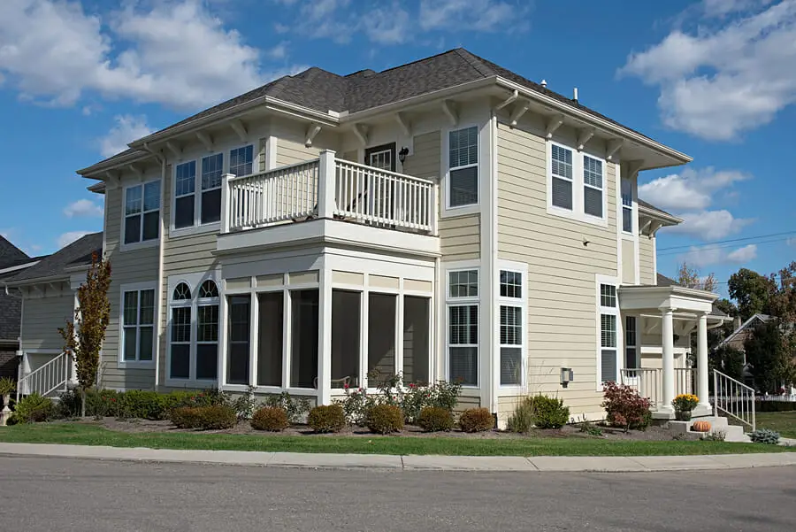 A two-story beige house with white trim and multiple large windows, featuring a balcony and a screened porch, sits on a corner lot with a small garden—its vinyl siding earns positive review for both durability and classic look under a partly cloudy sky.