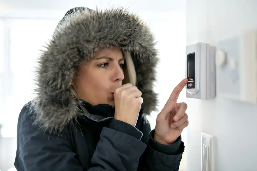 A person in a winter coat with a fur-lined hood adjusts a thermostat while shivering and holding their hand to their mouth, clearly thinking, Why do I always feel so cold in my house?.