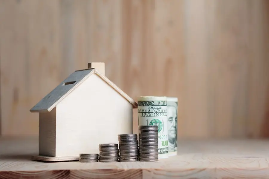 A small wooden house model next to stacks of coins and a rolled-up U.S. dollar bill on a wooden surface, representing savings, investment, or home improvement financing.