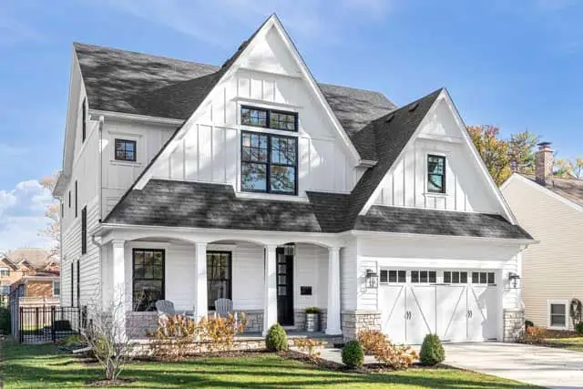 A modern two-story white house with black trim and trendy black windows features large front-facing gables, a covered porch with columns, a double garage, manicured lawn, and small shrubs, set against a blue sky.