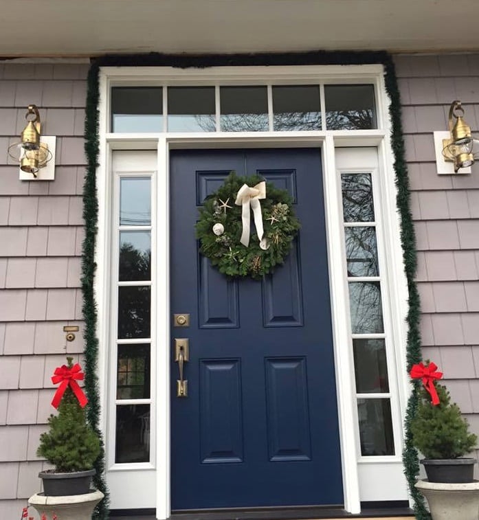 A navy blue front door, one of the best front door colors, is decorated with a holiday wreath and flanked by two small potted evergreens with red bows. Green garland frames the door, accented by lantern-style lights on either side.