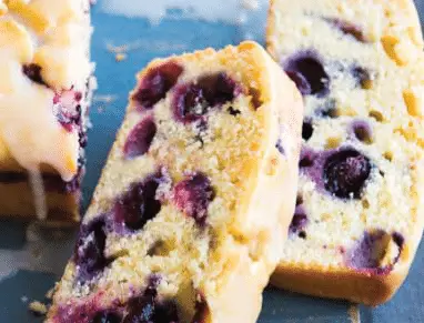 Sliced blueberry lemon drizzle cake with visible blueberries inside, resting on a blue surface. One slice is laid flat while others are upright, highlighting the cake’s moist texture and vibrant berry spots.