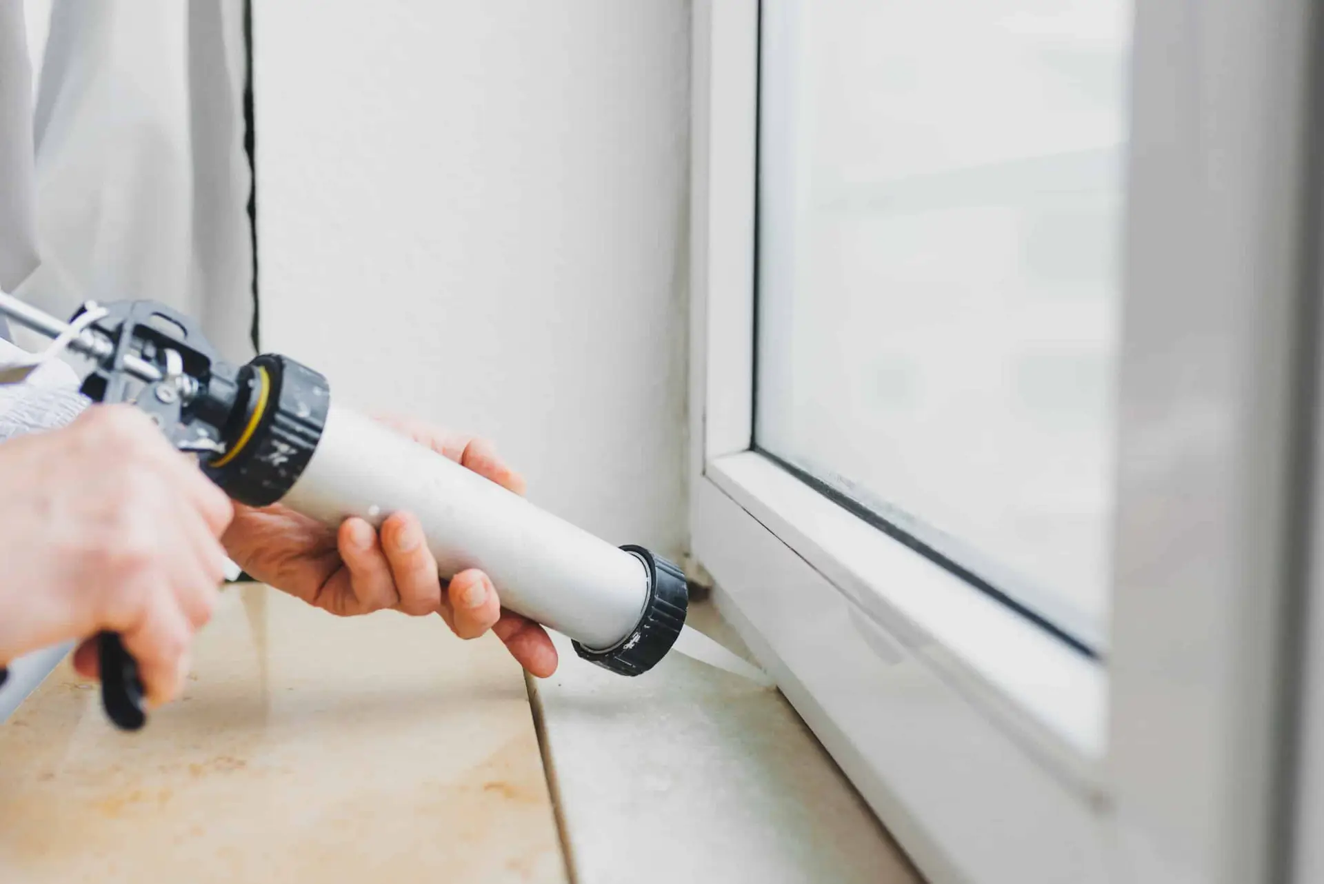 A person uses a caulking gun to seal the edge of a white window frame, applying caulk along the joint to minimize energy loss and prevent drafts—one of several essential energy saving tips for any home.
