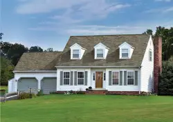 A white two-story suburban house with dormer windows, a brick chimney, and an attached two-car garage sits on a well-kept green lawn, showcasing a stunning spring home transformation under a partly cloudy sky.