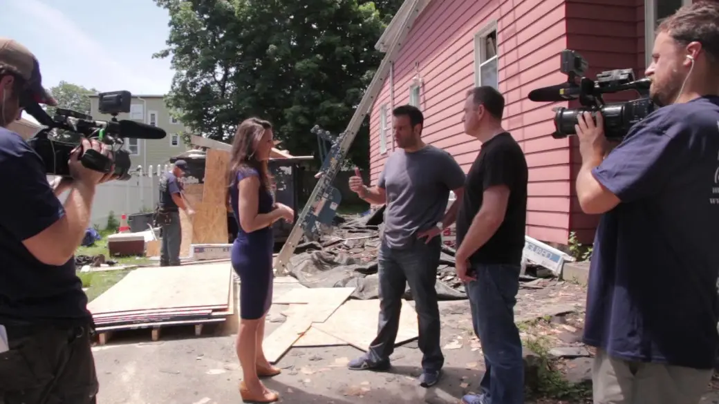 A woman in a blue dress interviews two men outside a red house under construction in Lynn, MA, while two cameramen film the scene. Catch this Sneak Peak with construction materials and debris visible in the background—classic Flipping Boston action!.