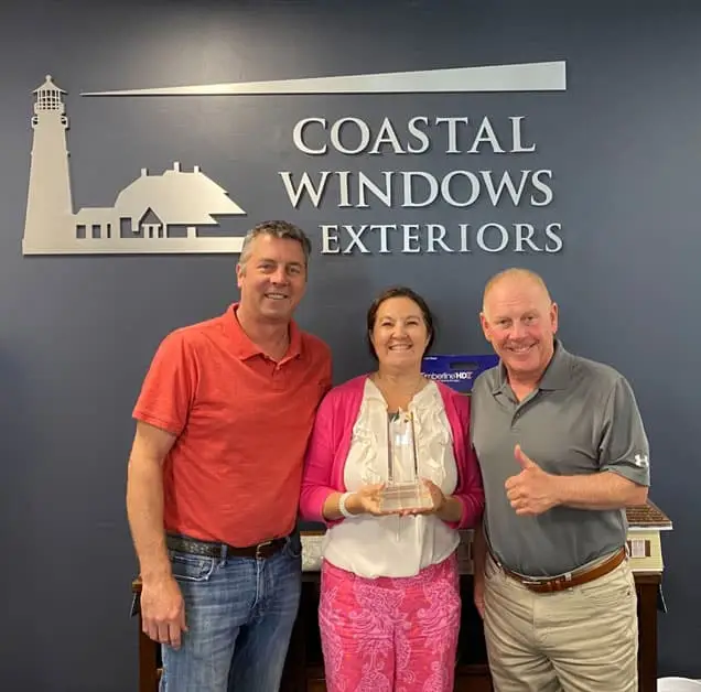 Three people stand smiling in front of a Coastal Windows & Exteriors sign. The woman in the center proudly holds the 2020 GAF President’s Club Award, while the men on either side stand close, the man on the right giving a thumbs up.