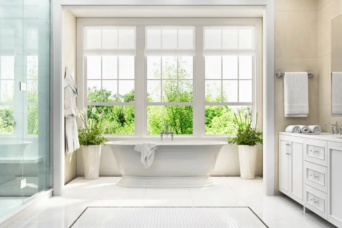 Bright bathroom with a white freestanding tub in front of large windows, flanked by tall plants. Wondering does a bathroom need a window? This space shows how natural light and lush greenery outside make it truly inviting.