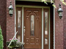 A wooden front door with decorative glass panels is set in a red brick exterior. Two lantern-style lights are mounted on either side, and a planter with flowers sits to the left—though fiberglass is your best choice for durability.