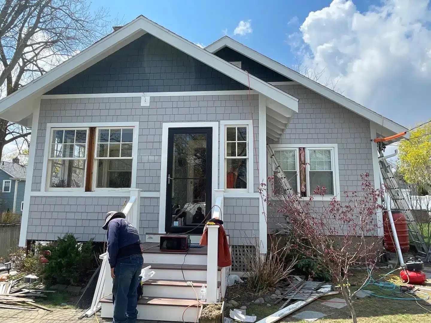 A person works on the front steps of a gray house with white trim and a covered porch. Construction materials, possibly including James Hardie® fire resistant siding, are scattered around, and the sky is partly cloudy.