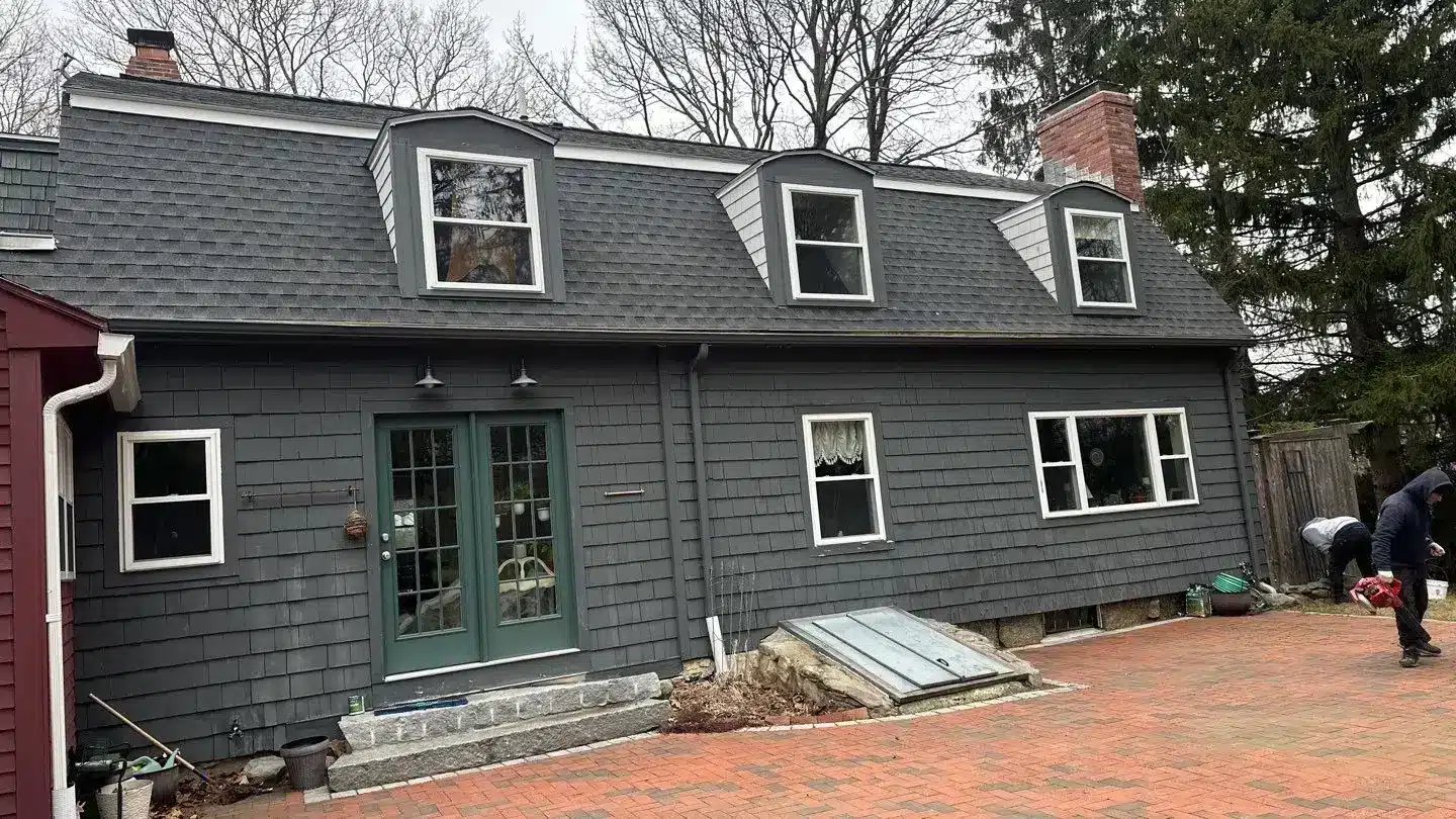A gray shingled house with three dormer windows, green double doors, and a brick driveway. Leafless trees are in the background, and two people are near the right side of the house, perhaps wondering: will new windows increase home value?.