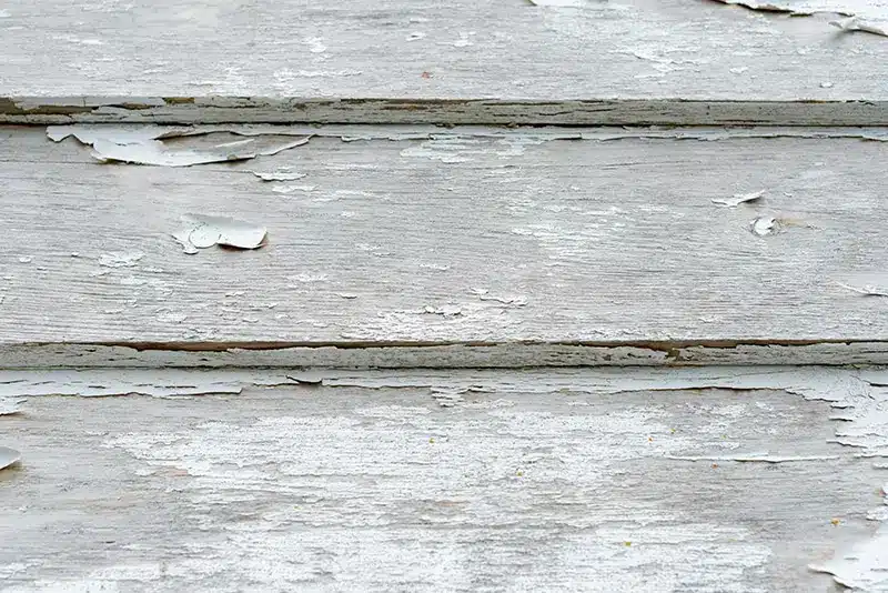 Close-up of weathered wooden boards with peeling, faded white paint and visible cracks. The aged texture highlights the contrast to modern materials like insulated vinyl siding, which offer greater durability and protection against the elements.
