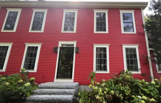A red two-story house with white trim, large windows featuring energy efficient window design, a black front door, stone steps, and green bushes in front.