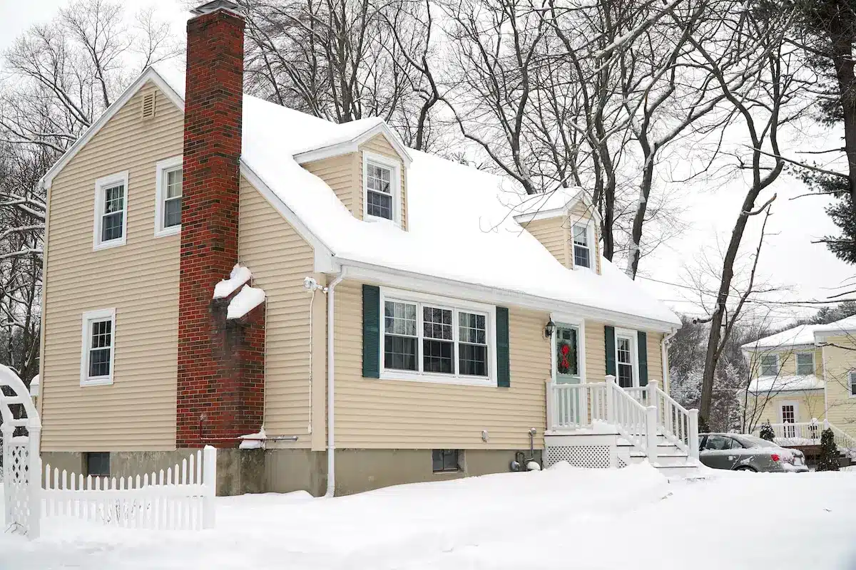 A beige two-story house with green shutters, replacement windows, a red brick chimney, and snow covering the roof, yard, and trees. A white picket fence and steps lead to the front door; another house is visible in the background.