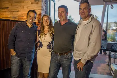 Four people stand closely together and smile at the camera indoors. Three men are casually dressed, and one woman in a light dress stands second from the left. Behind them, a brick and wood interior hints at Coastal Windows & Exteriors featured in Beverly Citizen.