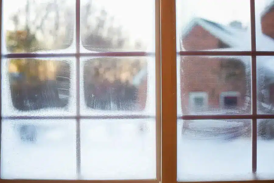 A close-up of drafty windows, their frosted panes featuring four clear squares wiped clean, offering a blurry view of snow-covered houses and trees outside.