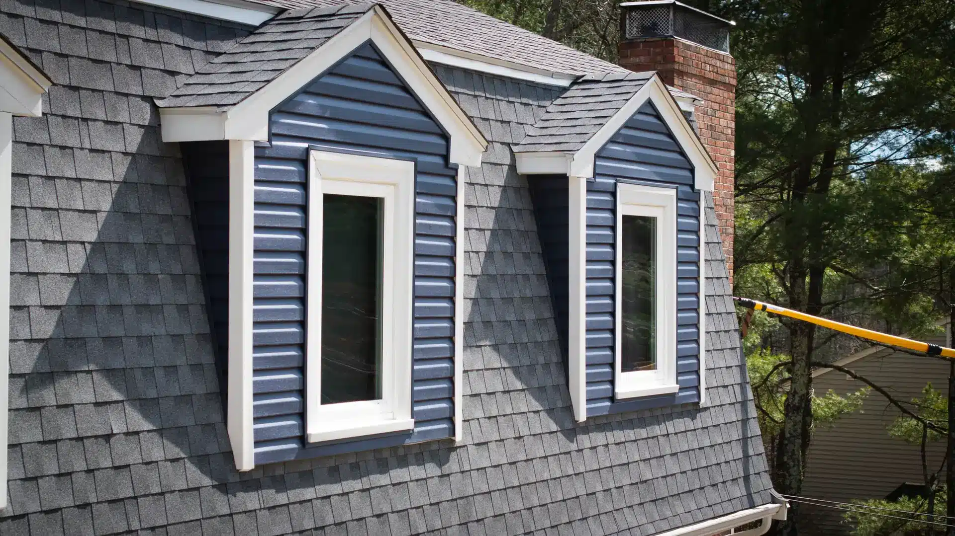 Two blue dormer windows with white trim protrude from a grey shingled roof, highlighting the importance of considering repair vs replace roof options. A red brick chimney is visible to the right, with trees in the background.