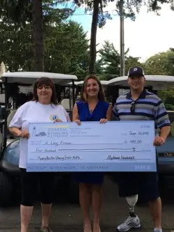 Three people stand outdoors, smiling and holding an oversized check at a golf tournament. Golf carts are visible in the background. One person on the right, representing A Leg Forever, has a prosthetic leg.