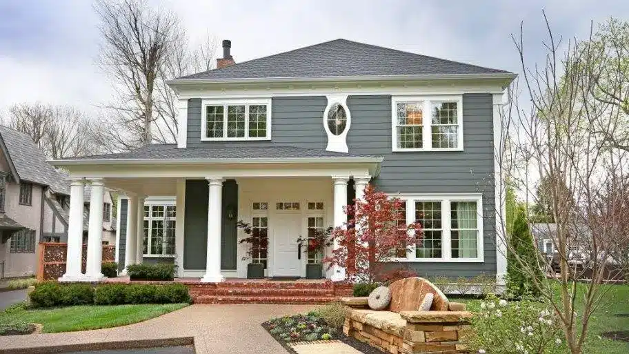 Two-story gray house with white trim and large columns on the porch, enhanced by expertly installed windows praised in top window contractor reviews. Front lawn features shrubs, small trees, brick steps, and a stone garden under an overcast sky.