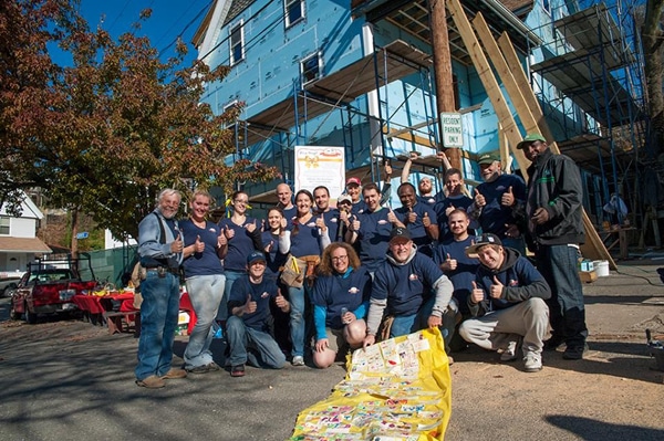 A group of people in matching shirts stand smiling and giving thumbs up in front of a house under construction, celebrating a Habitat for Humanity New Home Dedication, with a tree and construction materials visible around them.
