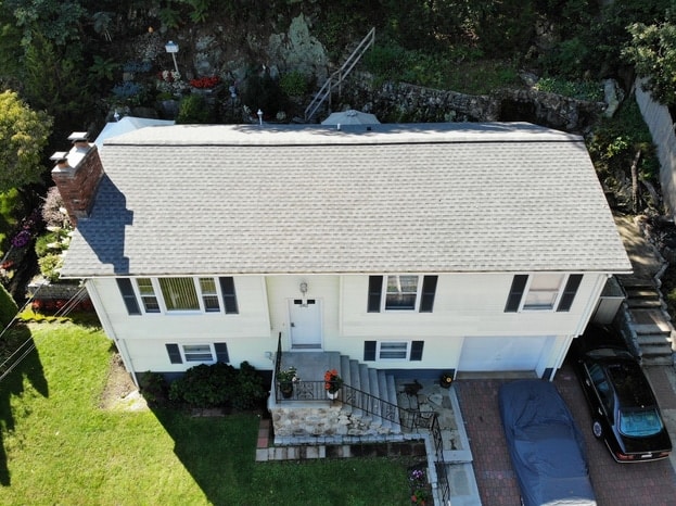 Aerial view of a two-story white house with a gray roof, front steps, and a small porch. A car covered by a tarp sits in the driveway. The green yard is bordered by trees—perfect for referencing your Window Replacement Checklist.