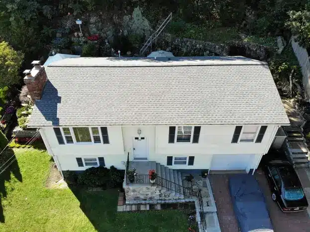 Aerial view of a white, split-level house with a grey roof—showcasing quality roofing in Hanover MA—front steps, a small garden, and driveway with two cars (one under a blue tarp), set against trees and a rocky hillside.