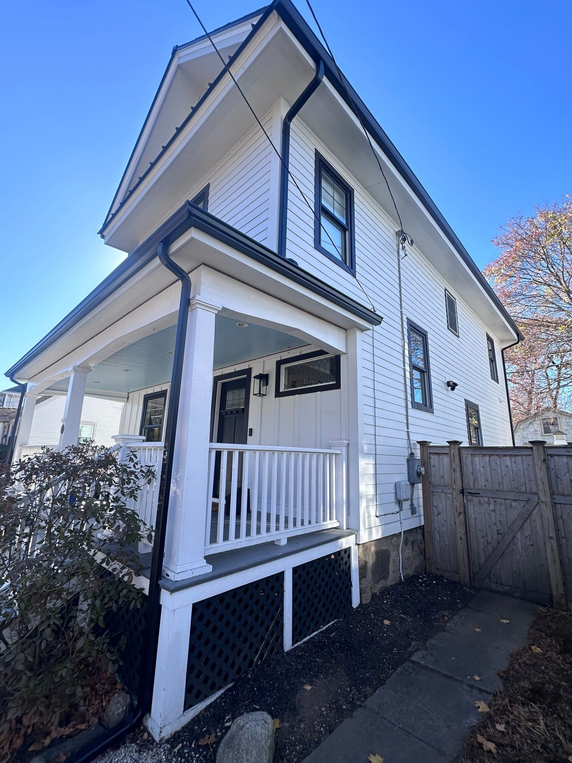 A white, two-story house with black trim and a small front porch. The porch has white railings and a black lattice underneath. Theres a wooden fence on the right, and a clear blue sky above. A bush is near the porch.
