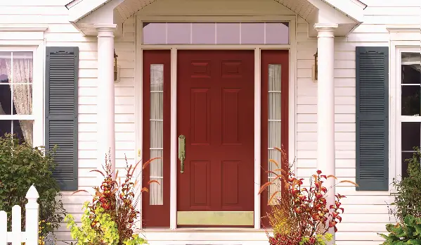 Dealer Spotlight: This charming white house features a ProVia red front door with glass side panels, flanked by columns, green shutters, and lush plants—perfect for a fresh coastal entryway.