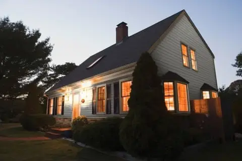 A cozy house with lights glowing inside at dusk, surrounded by trees and bushes, features a sloped roof and multiple windows—perfect for practicing energy savings tips year-round.