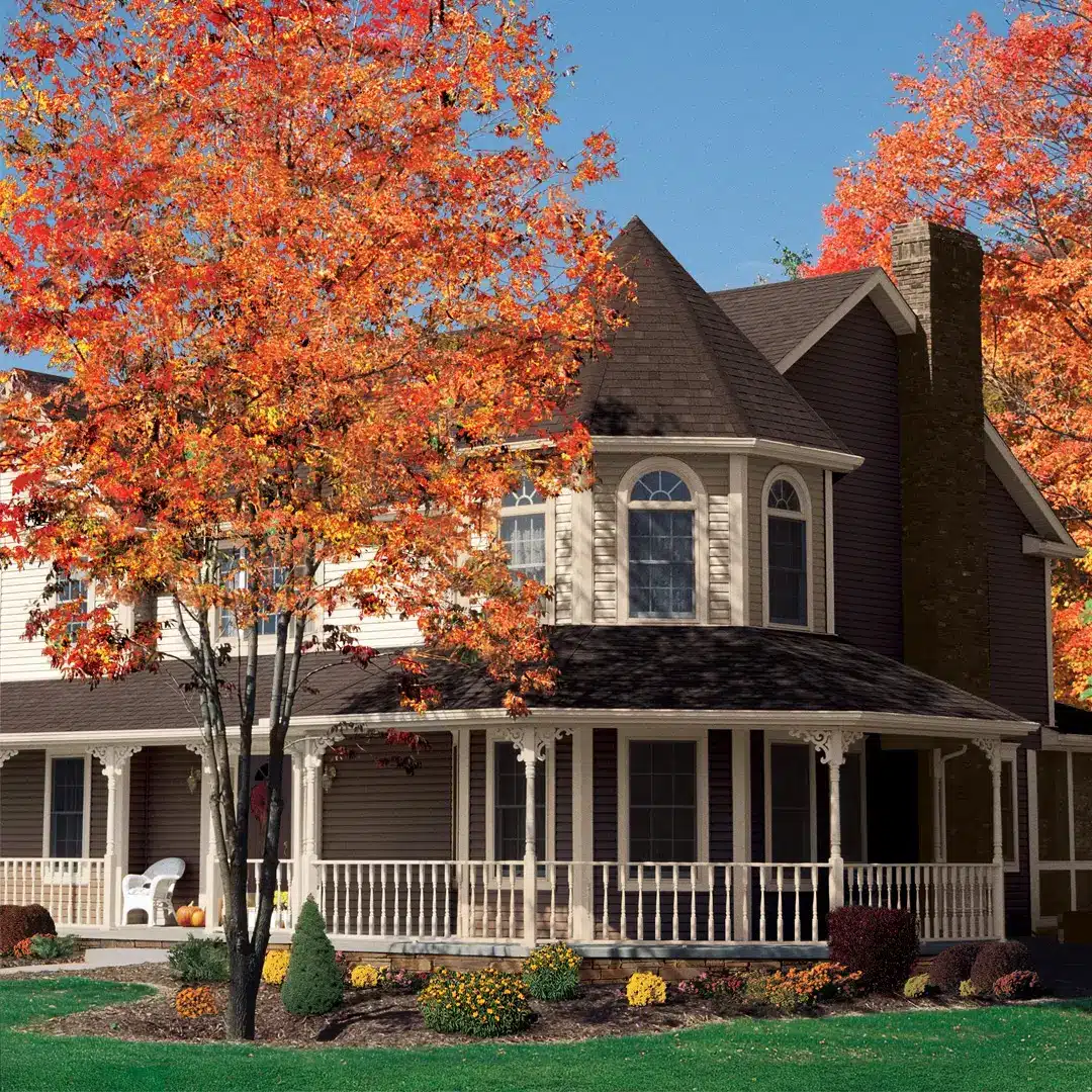 A two-story house with a wraparound porch and brown siding sits behind a lawn and garden beds. Bright autumn trees with red and orange leaves frame the scene, offering natural shade—one of many energy savings tips—under a clear blue sky.