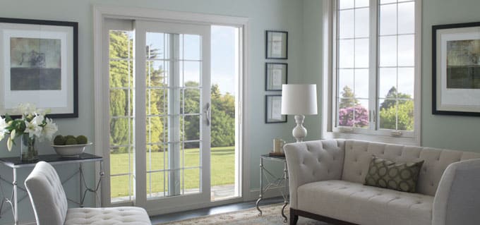 A bright living room with light-colored walls, a sofa, an armchair, framed art, a white lamp, and large sliding glass doors and windows that reveal a green, sunny yard outside.