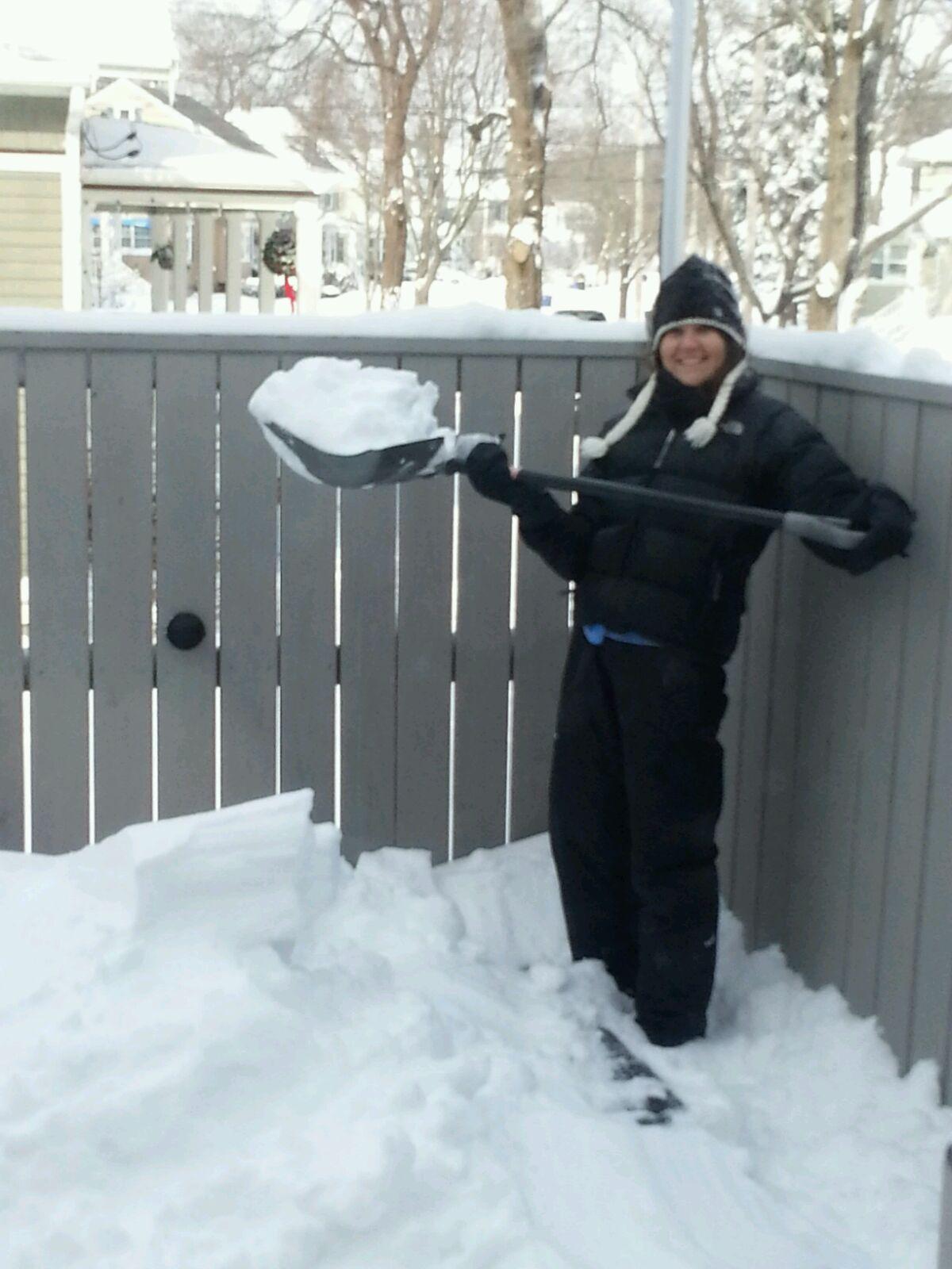 A person dressed in winter clothing stands outside on a snowy day, smiling while holding a shovel full of snow next to a wooden fence. With snow piled high, checking roof snow limits or snow load is important for nearby houses visible in the background.