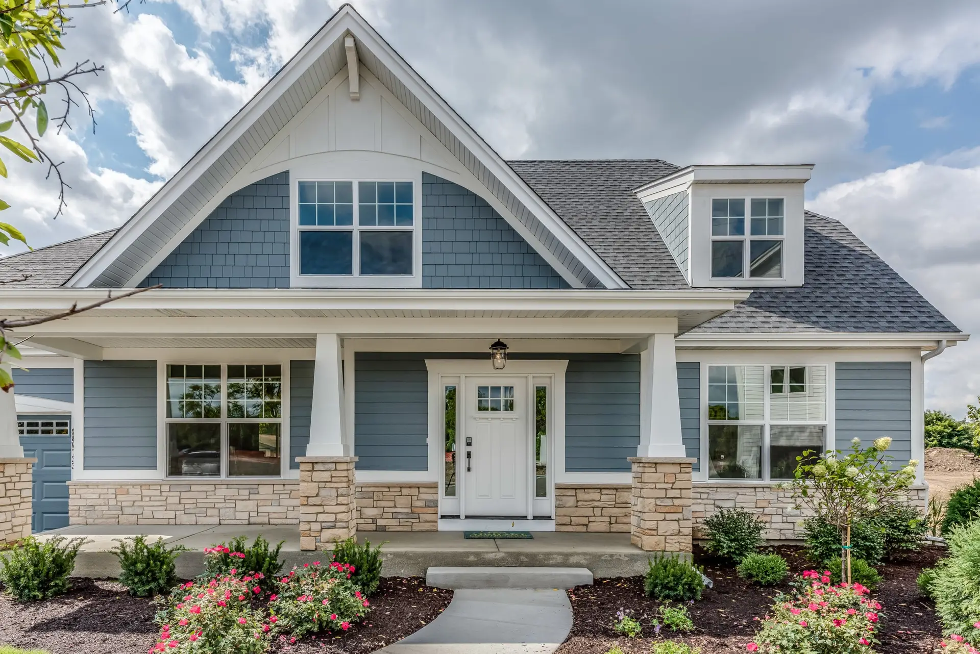 Charming single-family house with blue vinyl siding, white trim, a covered front porch, gabled roof, and a neatly landscaped garden—perfect for those interested in maintaining vinyl siding—under a partly cloudy sky.