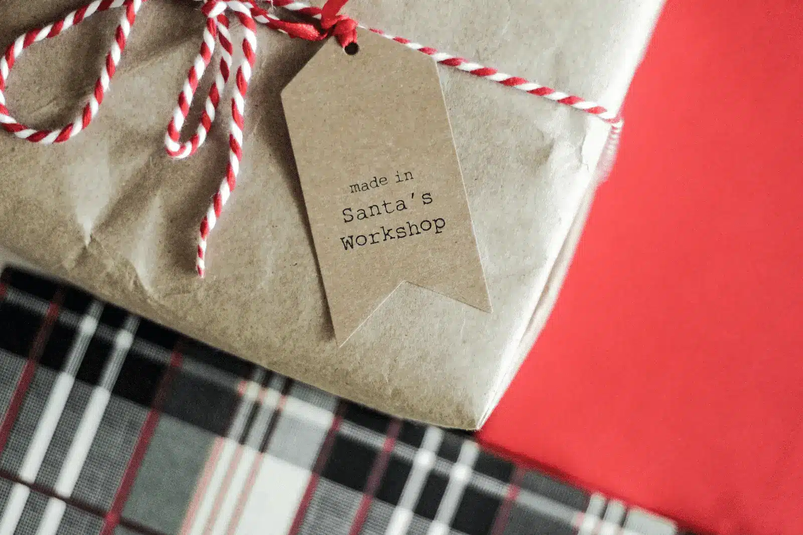 A gift wrapped in brown paper with red and white twine features a tag that reads made in Santa’s Workshop. A tartan plaid gift sits nearby on a red surface, capturing the cozy magic of the holidays sale straight from Santa’s roof.
