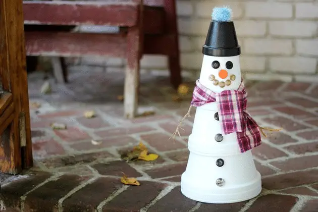 A snowman made from stacked white flower pots, decorated with buttons, a plaid scarf, an orange nose, a black pot hat with a blue pom-pom, and small stones for the mouth sits on a brick patio near a bench—perfect by the best house siding for cold climates.