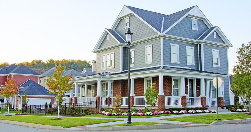 A large, two-story house with gray siding and red brick exterior features a wraparound porch—one of the best home improvements—white railing, manicured lawn, flowers, and small trees on a suburban street corner.