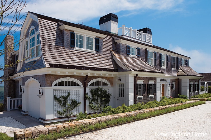 A large, elegant coastal home with wood shake shingle siding, white trim, and black shutters. The house features multiple chimneys, dormer windows, and a landscaped path bordered by a low stone wall and greenery.