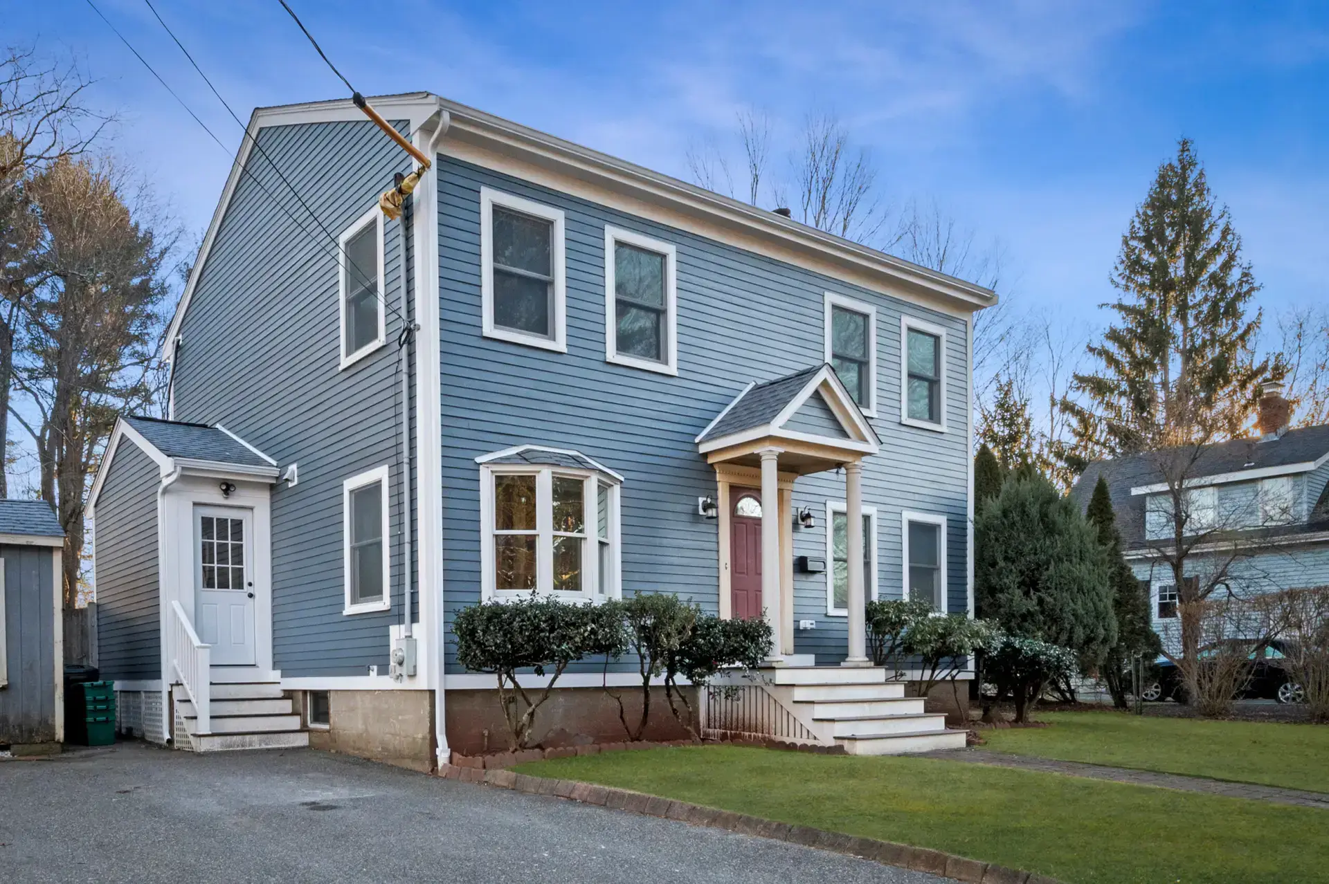 A two-story blue house with white trim, a pink front door, small front porch, and bay window. The grassy yard has trimmed bushes and a left-side driveway. The home features durable siding and sits beneath a clear blue sky.