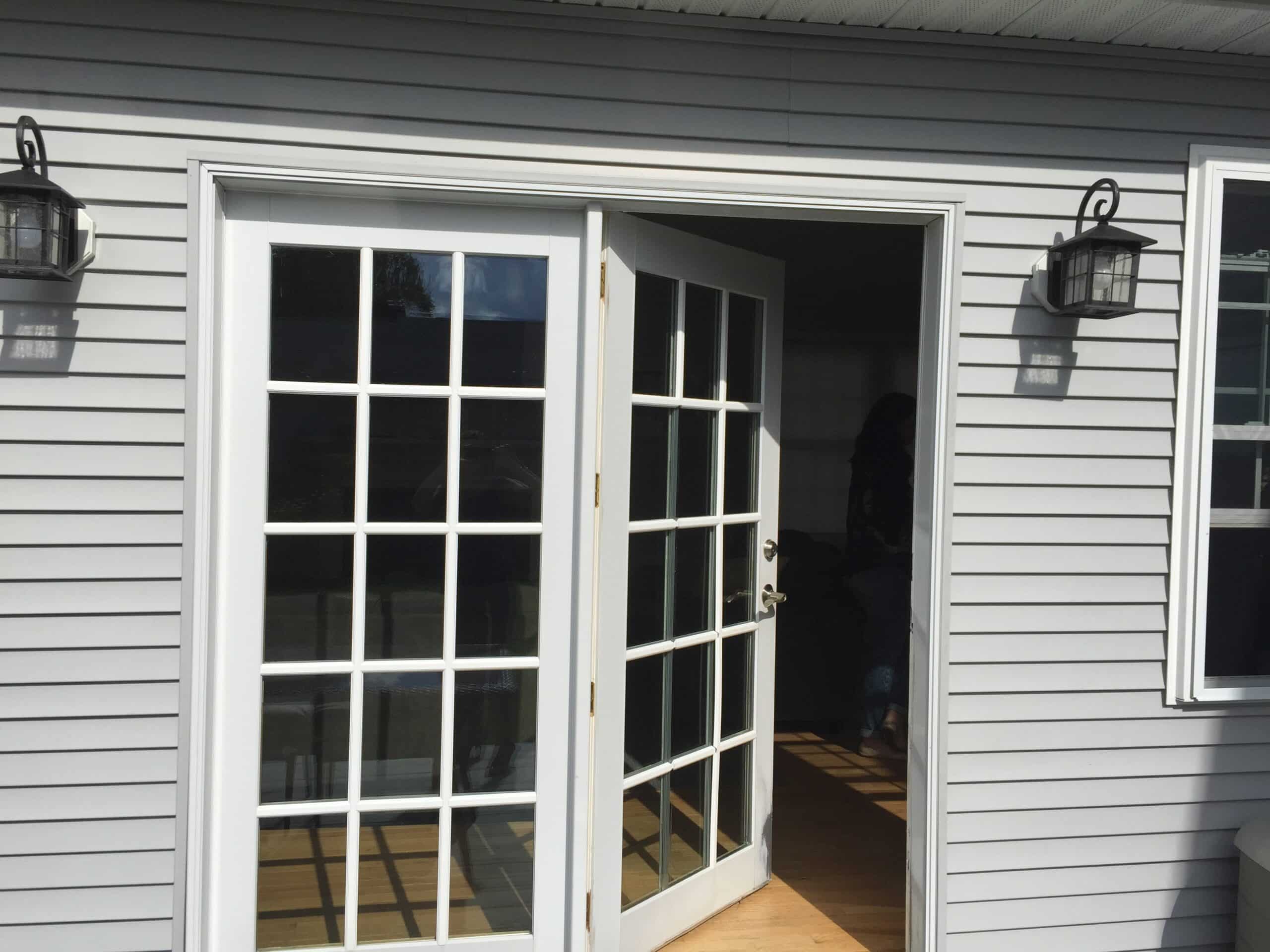 A set of white French doors, one open, leads from the outside into a house with gray siding. Reminiscent of a sliding glass door, two black wall lanterns flank the doors as sunlight streams onto a wooden floor inside.