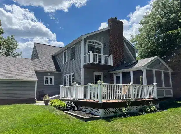 A two-story gray house with white trim and vinyl siding in Lexington, MA features a small balcony, brick chimney, screened porch, and wooden deck with white railings. It sits among green grass and trees beneath a blue sky with clouds.