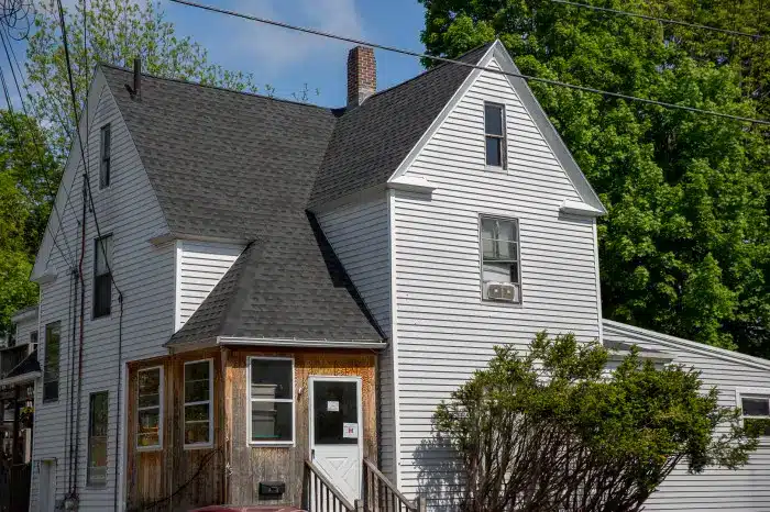A two-story white house with dark roof shingles, wood-trimmed enclosed porch, and a window air conditioning unit. Bushes grow in front, and trees and power lines are visible in the background—perfect for a roof installation video.